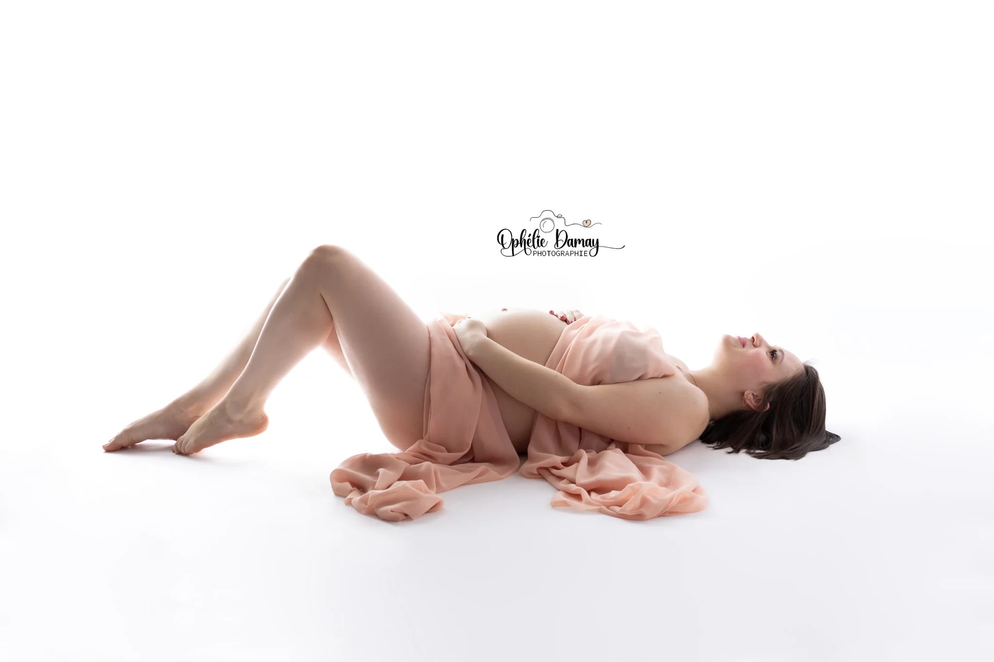 alt="Femme enceinte allongée sur le dos, lumière douce en studio"