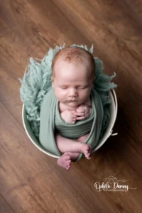 alt="Photo de nouveau-né emmailloté dans un panier, lumière douce en studio photo"