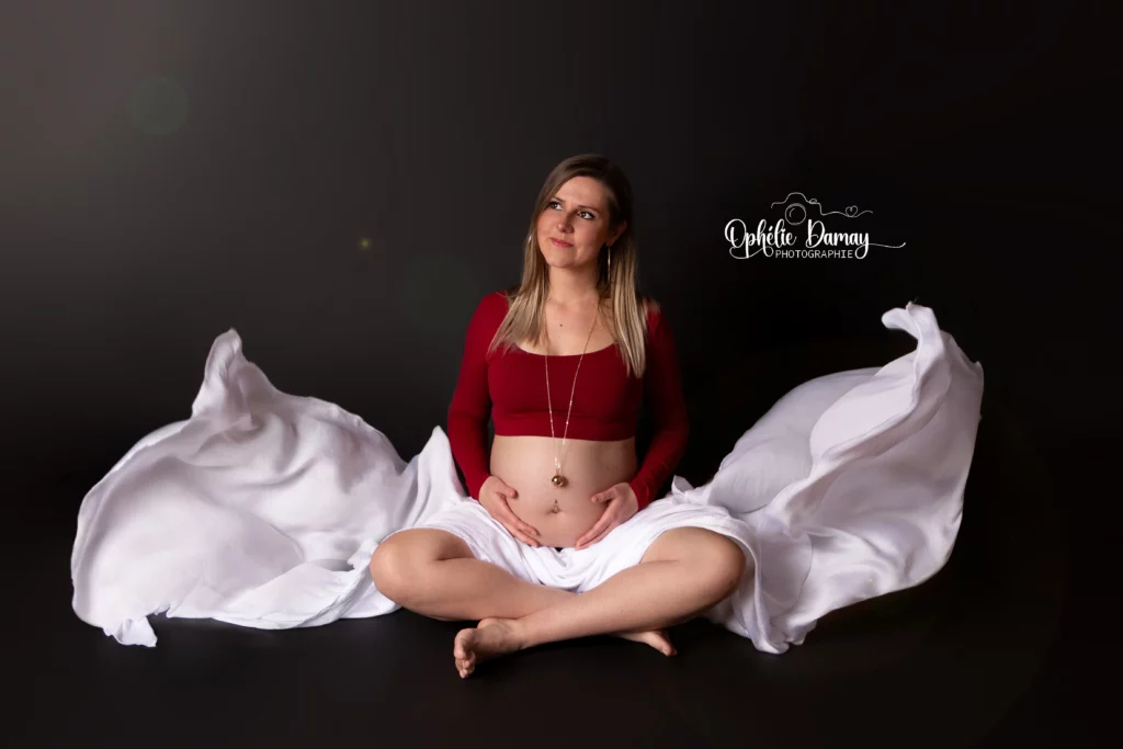alt="Femme enceinte, assise en tailleur avec un voilage qui vole de chaque côté d'elle"