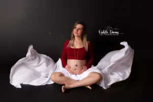 alt="Femme enceinte, assise en tailleur avec un voilage qui vole de chaque côté d'elle"