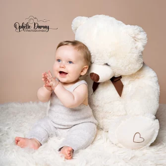 alt="Bébé assis sur un tapis de laine contre un ours en peluche, applaudissant vivement"