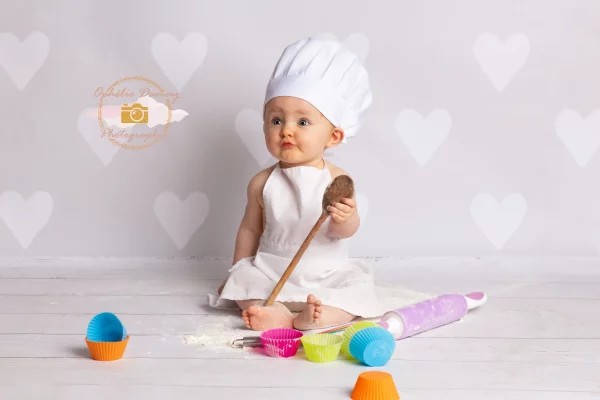 alt="Bébé habillé en cuisinier et entouré d'ustensiles de cuisine et de farine"